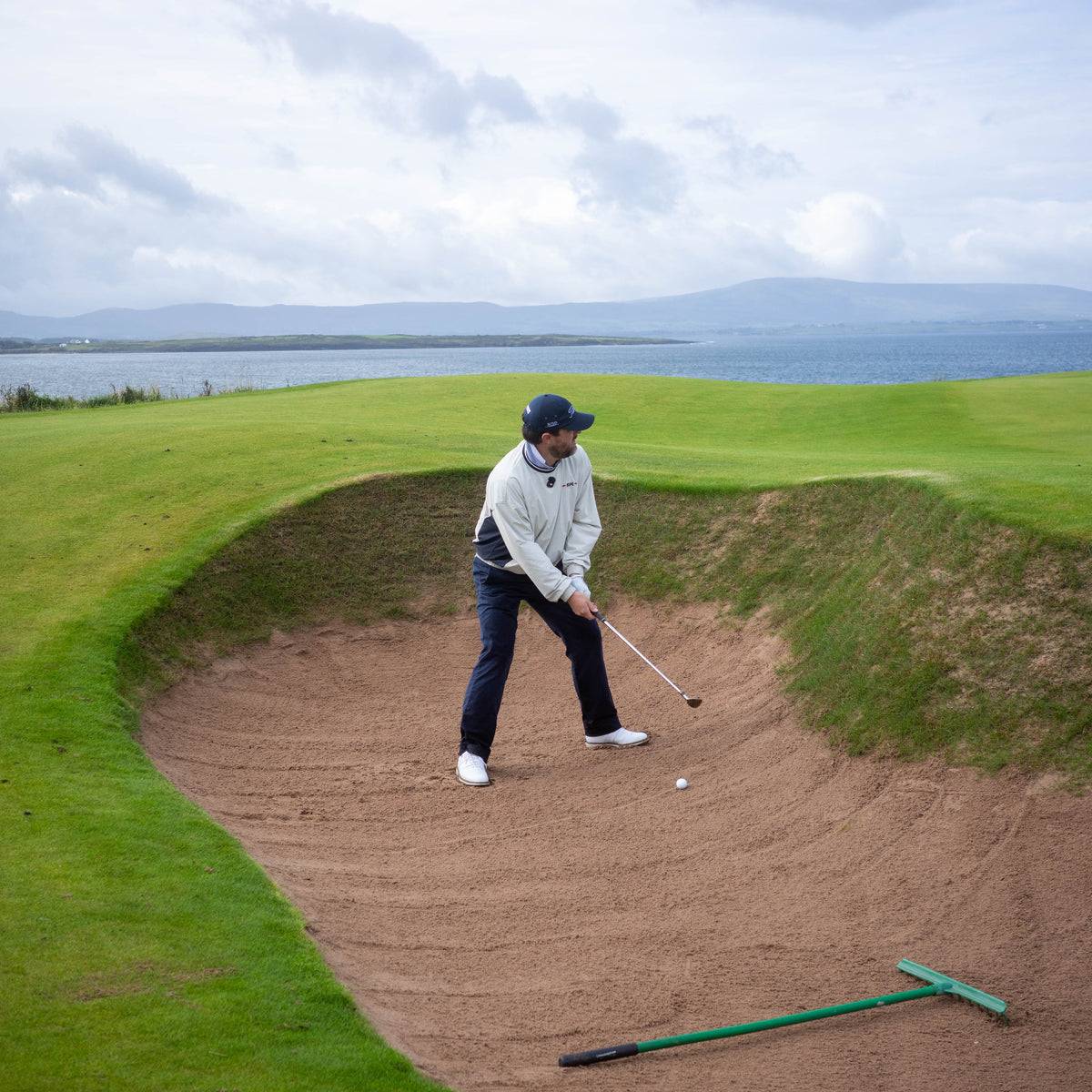 A golfer in a white jacket and dark pants stands in a sand bunker on a golf course, preparing to hit a golf ball with a club. A green rake lies nearby, with grass, water, and a cloudy sky in the background.