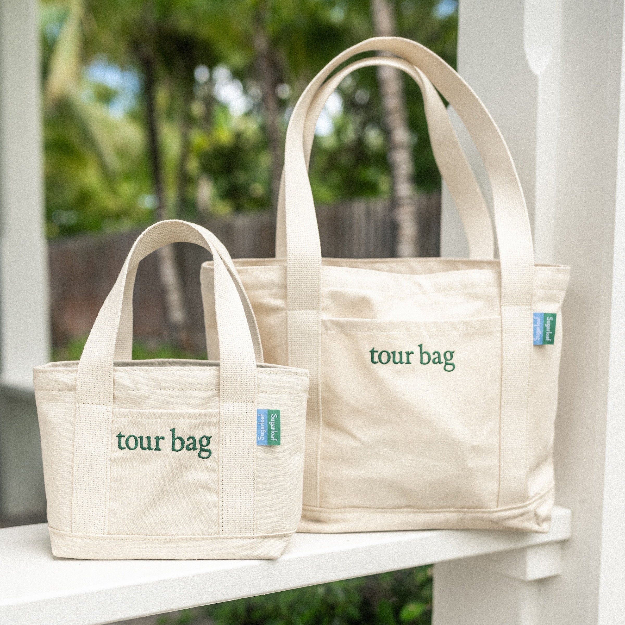 Two beige tote bags labeled 'tour bag' on a white surface with a blurred green outdoor background.