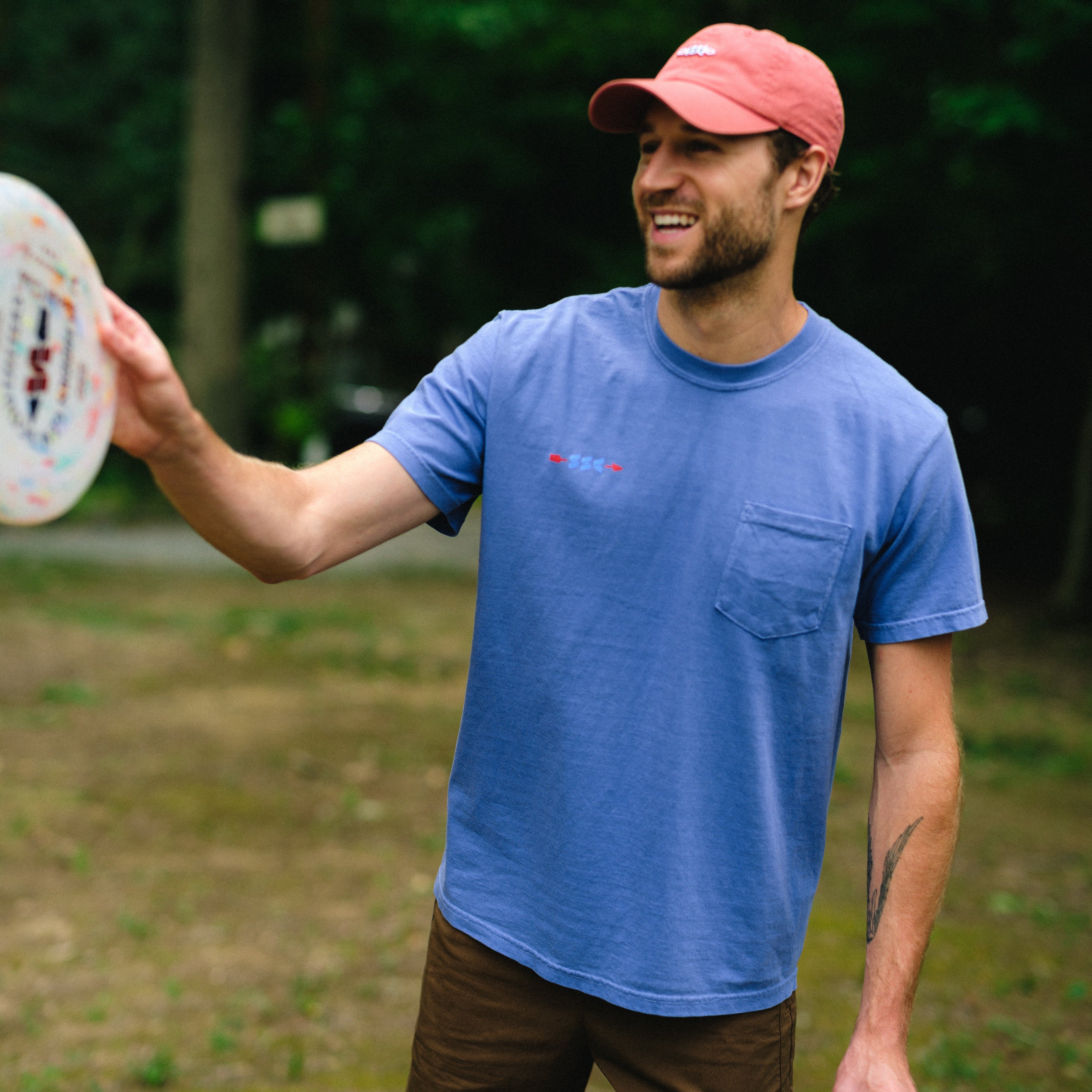 A man outdoors in a park wearing a blue crewneck pocket t-shirt, brown shorts, and a pink cap, holding a disc golf disc and a beverage can.