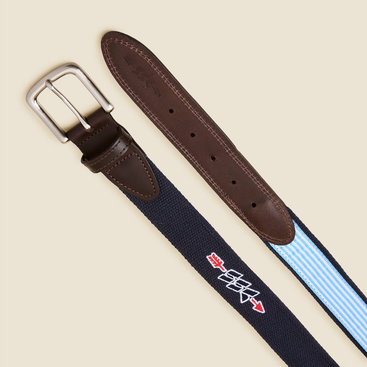 A close-up of a woven belt with a navy blue strap, light blue and white stripes on one end, dark brown leather end caps, and a silver buckle. The woven section features a white embroidered logo with red accents.