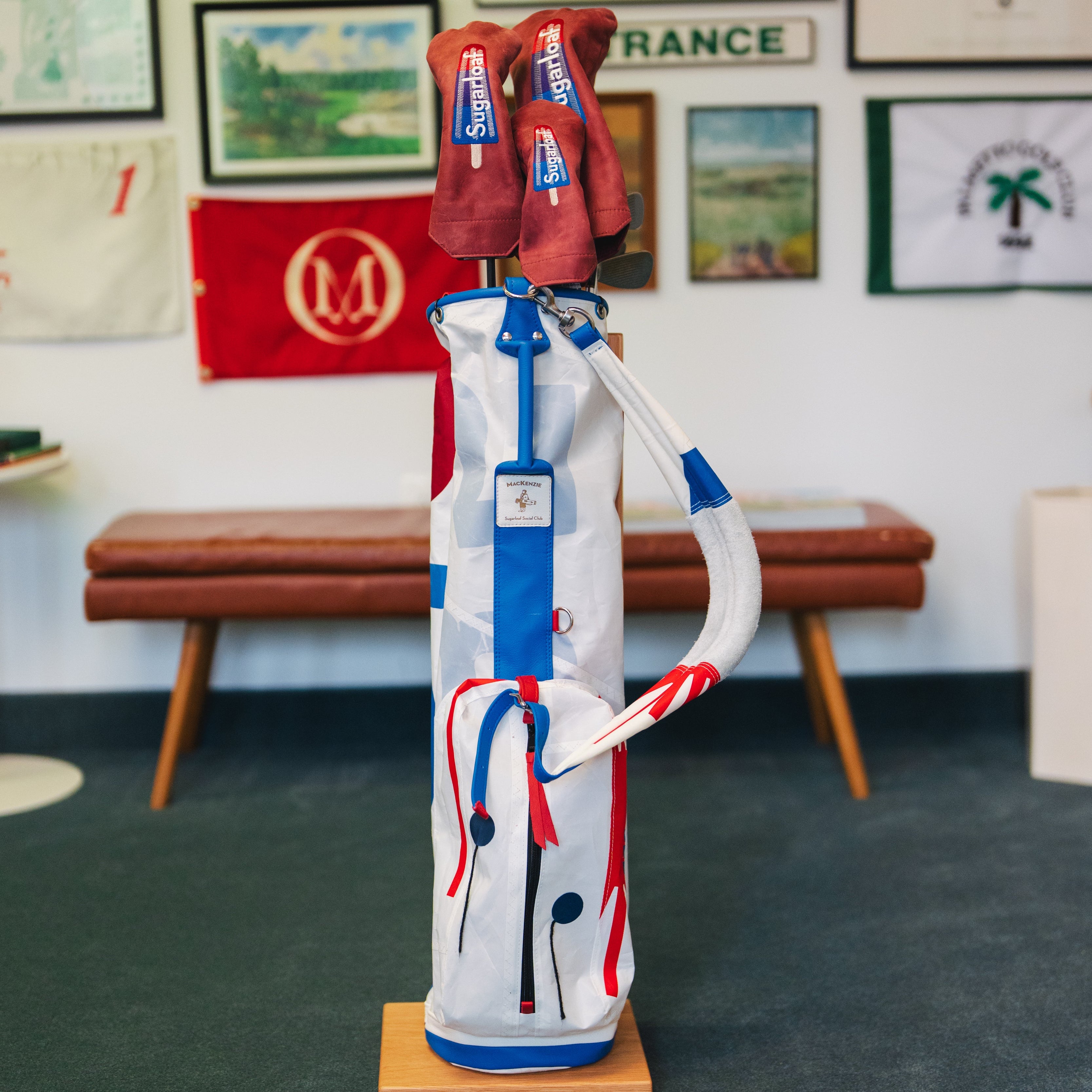 A golf club bag filled with red and blue golf clubs, displayed on a wooden stand in an indoor room with framed golf posters and a bench in the background.