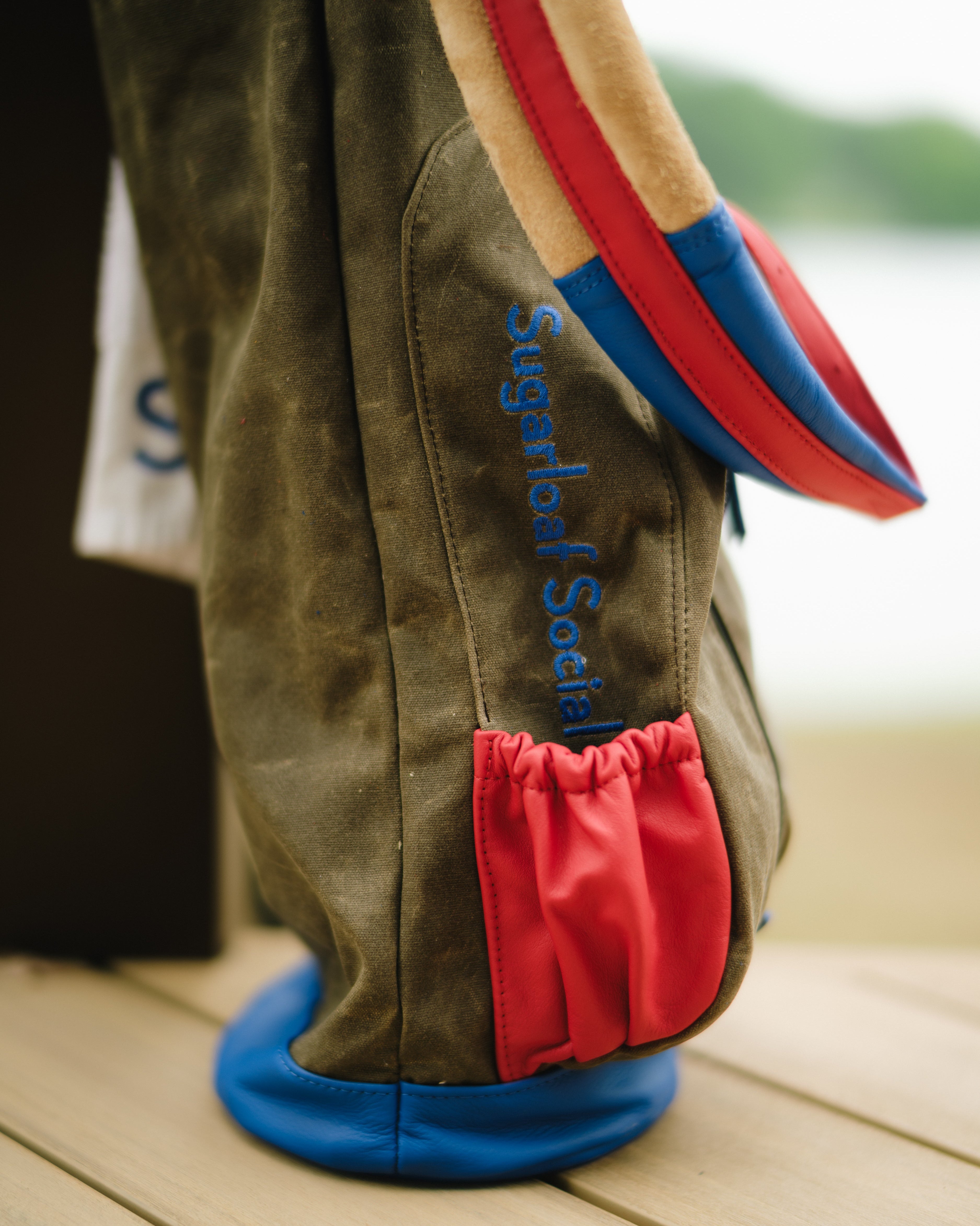A close-up of a suede golf bag standing on a wooden deck, with a blurred outdoor background. The bag is olive green with blue embroidered text reading 'Sugardahl Sockets', a red drawstring pocket, and red and blue trim.