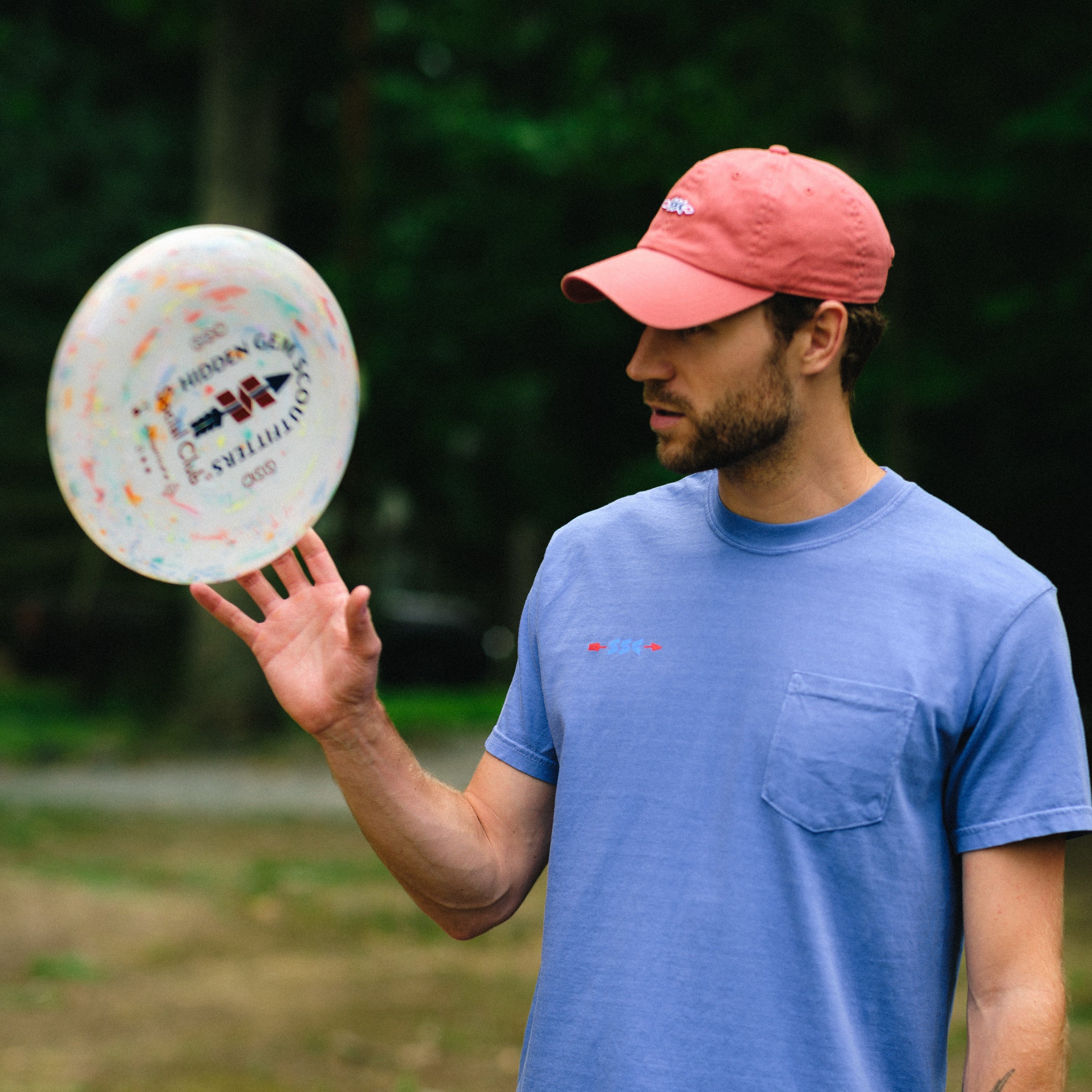 A man wearing a pink cap, a blue short-sleeve pocket t-shirt, and brown shorts stands outdoors in a wooded area, holding a white disc golf disc in one hand and a beer can in the other.
