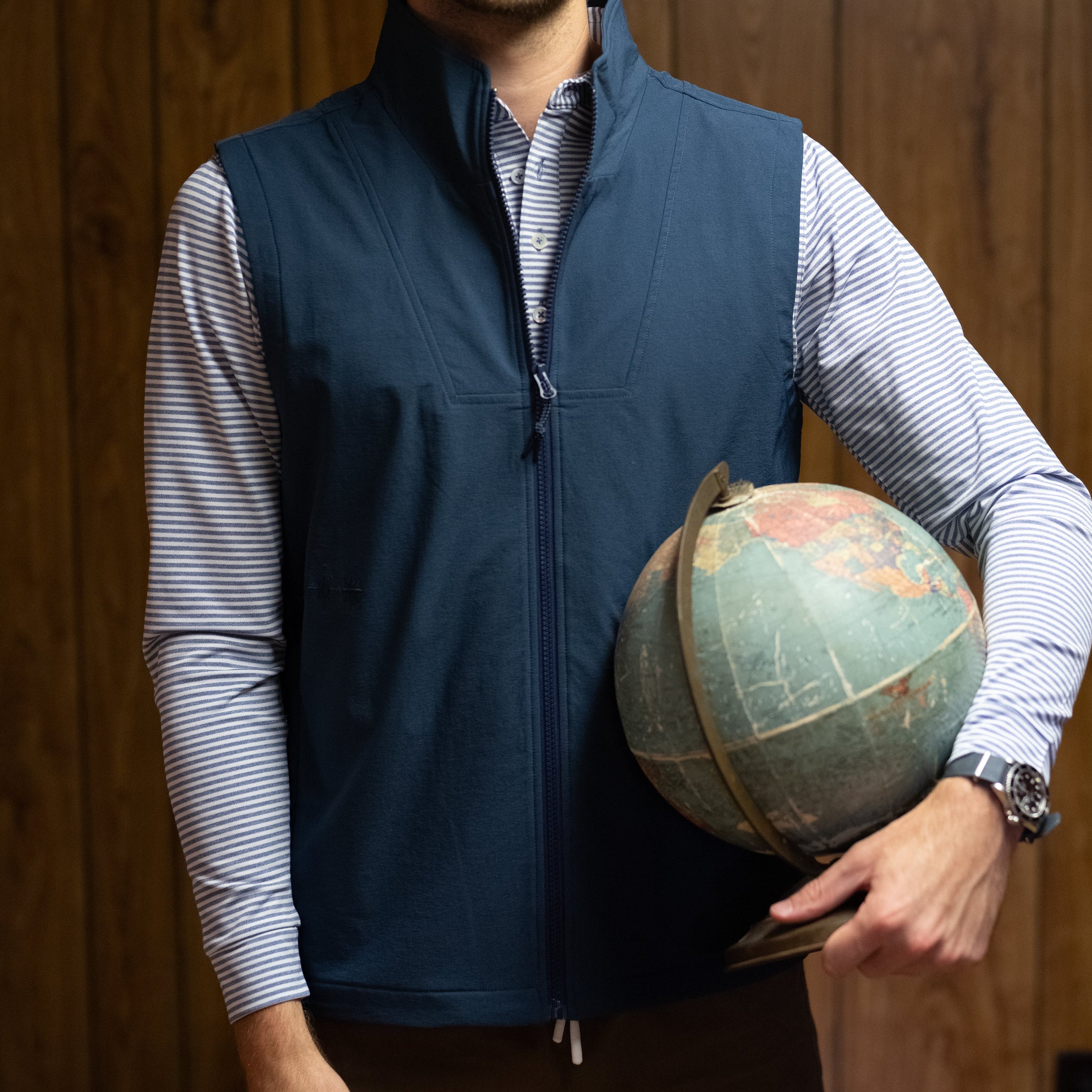 A man wearing a navy blue zip-up vest over a light blue and white striped long-sleeve shirt, holding a vintage globe in front of a wood-paneled wall.