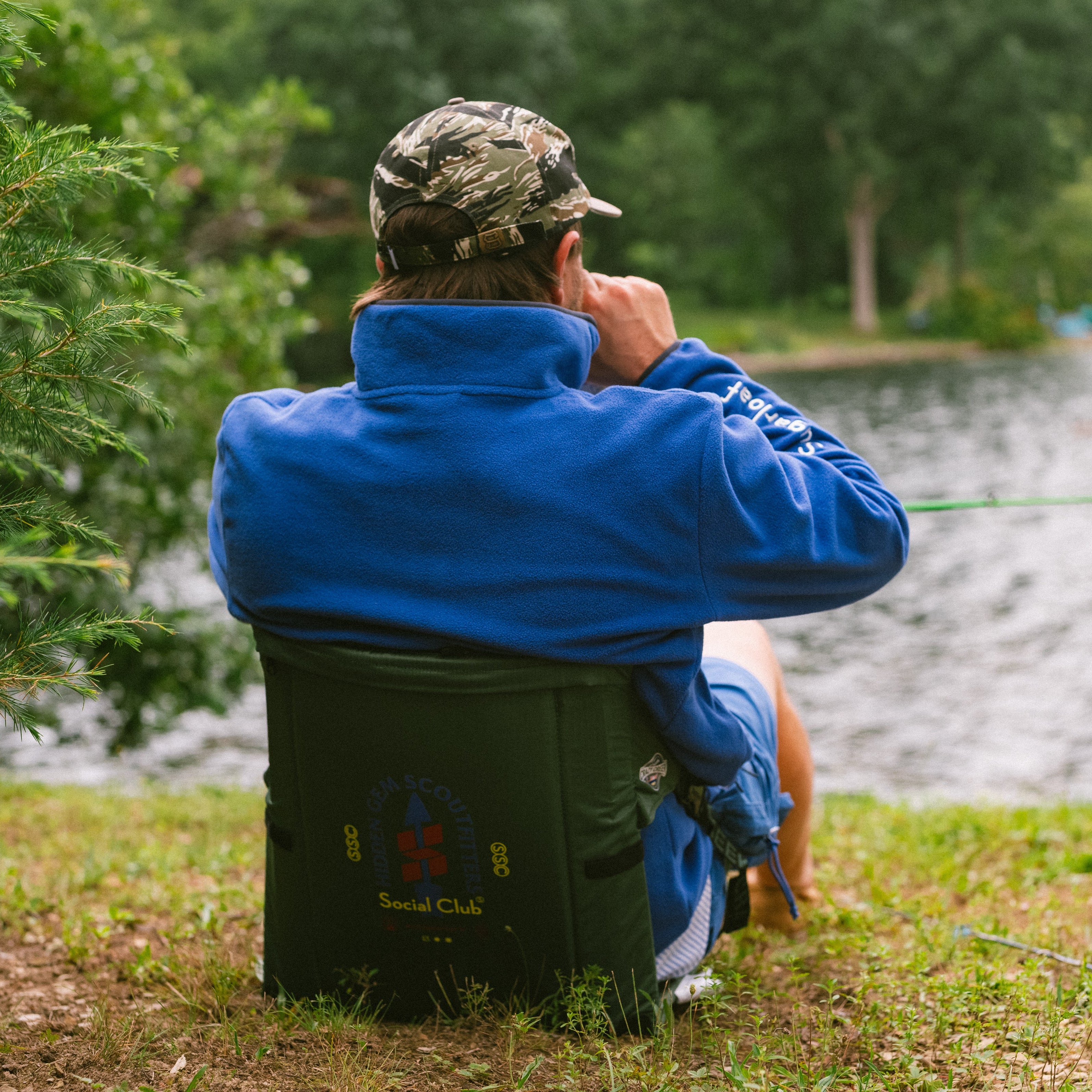 A man sits on a green fishing chair by a lake, viewed from behind as he looks out toward the water. He is wearing a blue jacket, a camouflage cap, and shorts, with a fishing rod resting on the grass nearby.