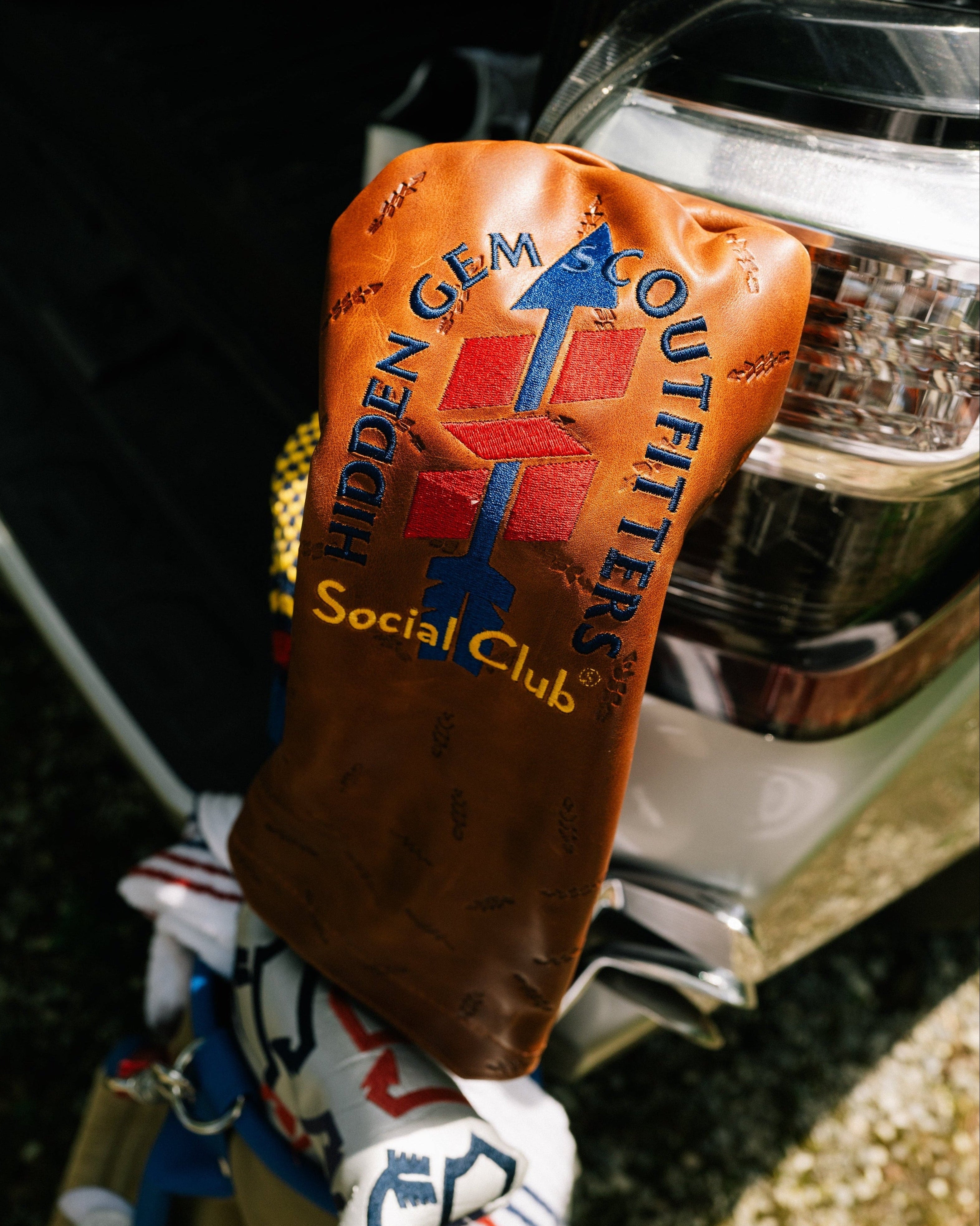 A brown leather golf club headcover with 'HIDDEN GEM CLOUTHERS Social Club' printed in blue and red, resting against a silver vehicle body.