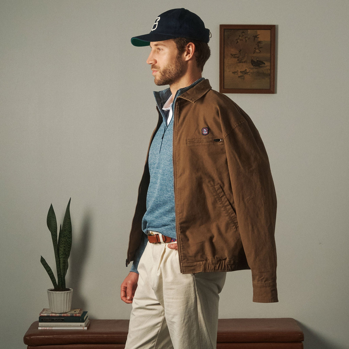 A man wearing a brown canvas utility jacket, light blue knit sweater, beige trousers, and a navy baseball cap stands in profile in a styled room with a bench and framed wall art.
